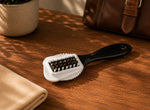 A 4-in-1 leather and shoe cleaning brush with a black handle, white bristle base, and brown bristles, resting on a wooden table with a folded beige cloth, a potted plant, and a brown leather bag in the background.