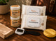 A product bundle on a wooden table featuring two jars of 'The Wonder Wax' natural hair wax, two packs of 'Peyton Instant Cleaning Pods', a beige towel, a black-handled brush, and a yellow sponge.